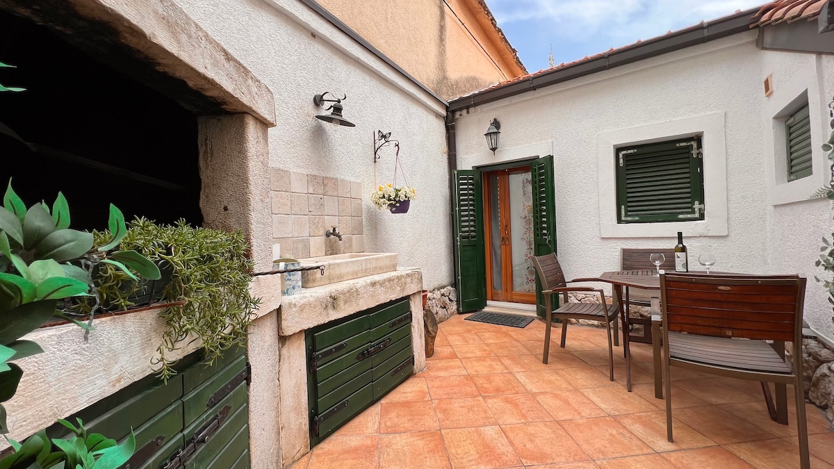 An outdoor patio area features a stone sink and a wooden dining table complemented by four chairs. Green shutters adorn the windows, adding a touch of charm. Potted plants provide a natural feel, while the terracotta tiles enhance the space's warm ambiance.
