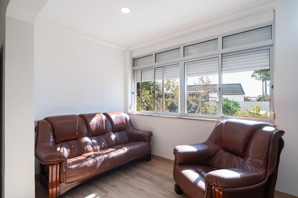 A light-filled sitting area is furnished with two brown leather sofas, positioned to maximize natural light from the large windows. The space features neutral-colored walls and wooden flooring, creating a calm atmosphere with views of the lush greenery outside.