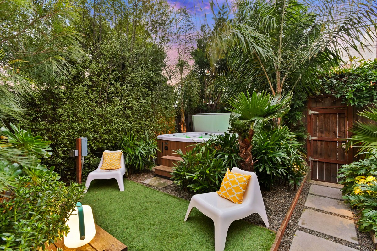 A tranquil garden area features lush greenery and palm plants, creating a serene atmosphere. Two modern chairs with yellow patterned cushions provide seating near a hot tub, which is nestled among the plants. A soft glow from a nearby lantern illuminates the space.