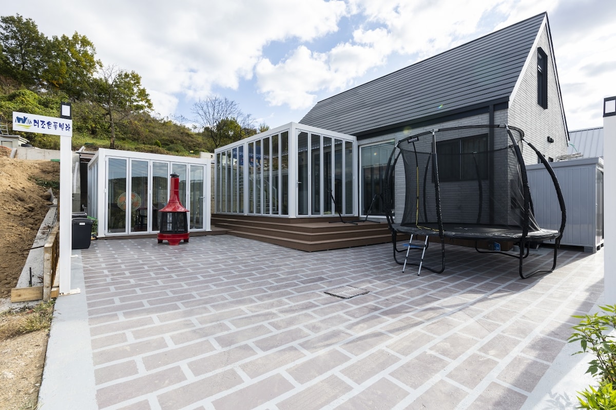 An outdoor area featuring a spacious patio with modern stone flooring. Large glass walls of the building allow natural light inside, while a trampoline and a red outdoor fireplace add recreational options to the space.