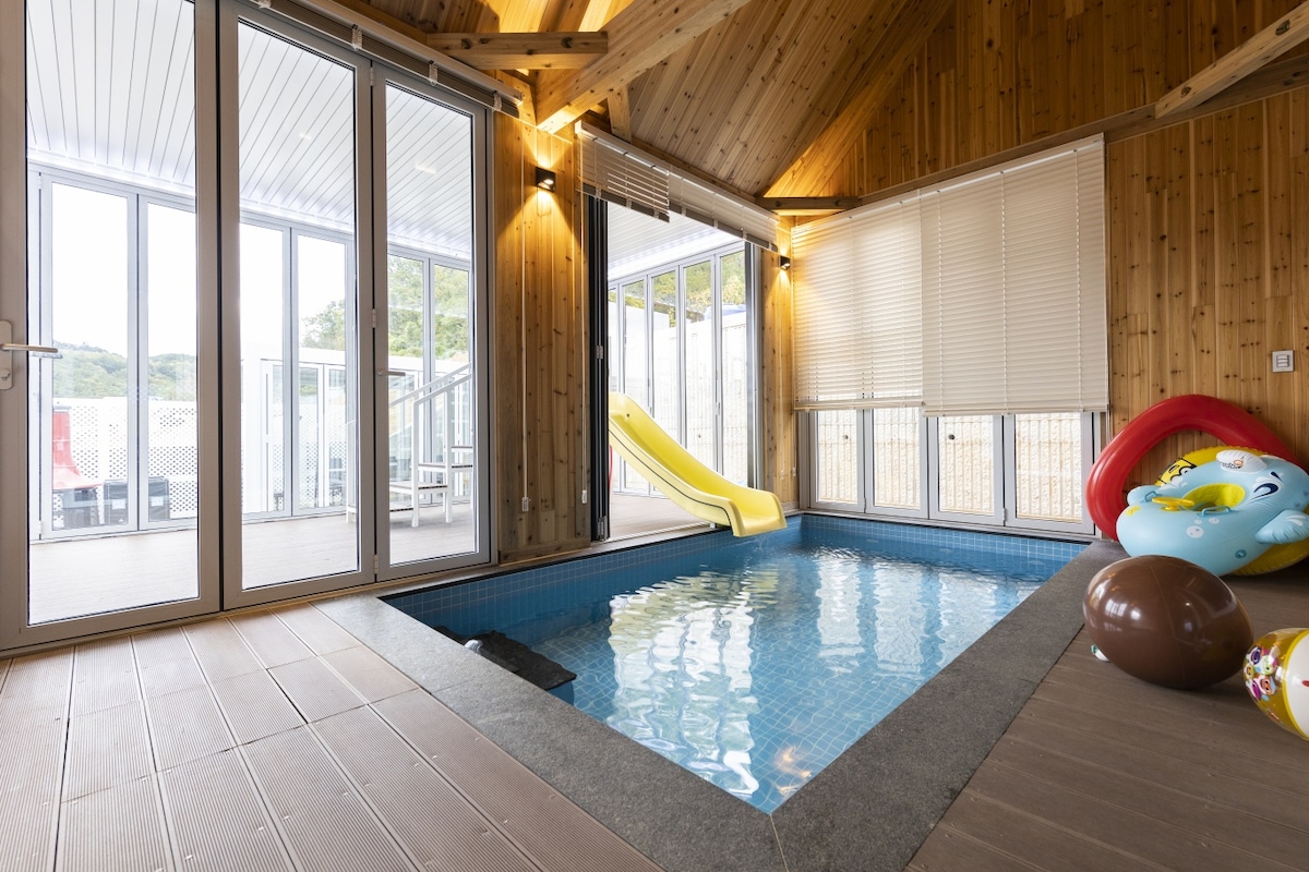 An indoor heated pool is framed by wooden walls and large glass doors, allowing natural light to fill the space. A slide leads into the water, complemented by colorful floating toys nearby. The area is designed for family fun and relaxation.