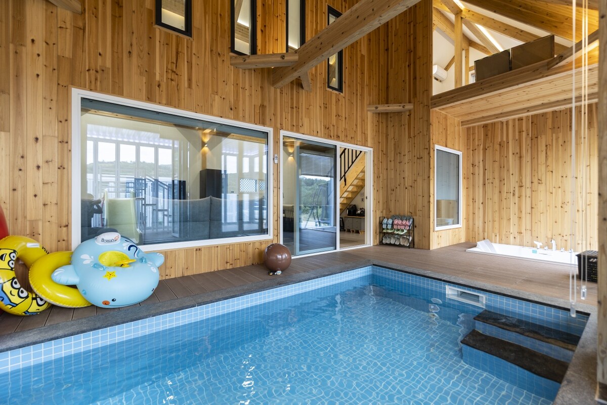 An indoor pool is featured within a wooden-paneled space, surrounded by large windows that allow natural light. Colorful inflatable toys rest beside the water, creating a playful atmosphere. Stairs lead to an upper level overlooking the pool area.