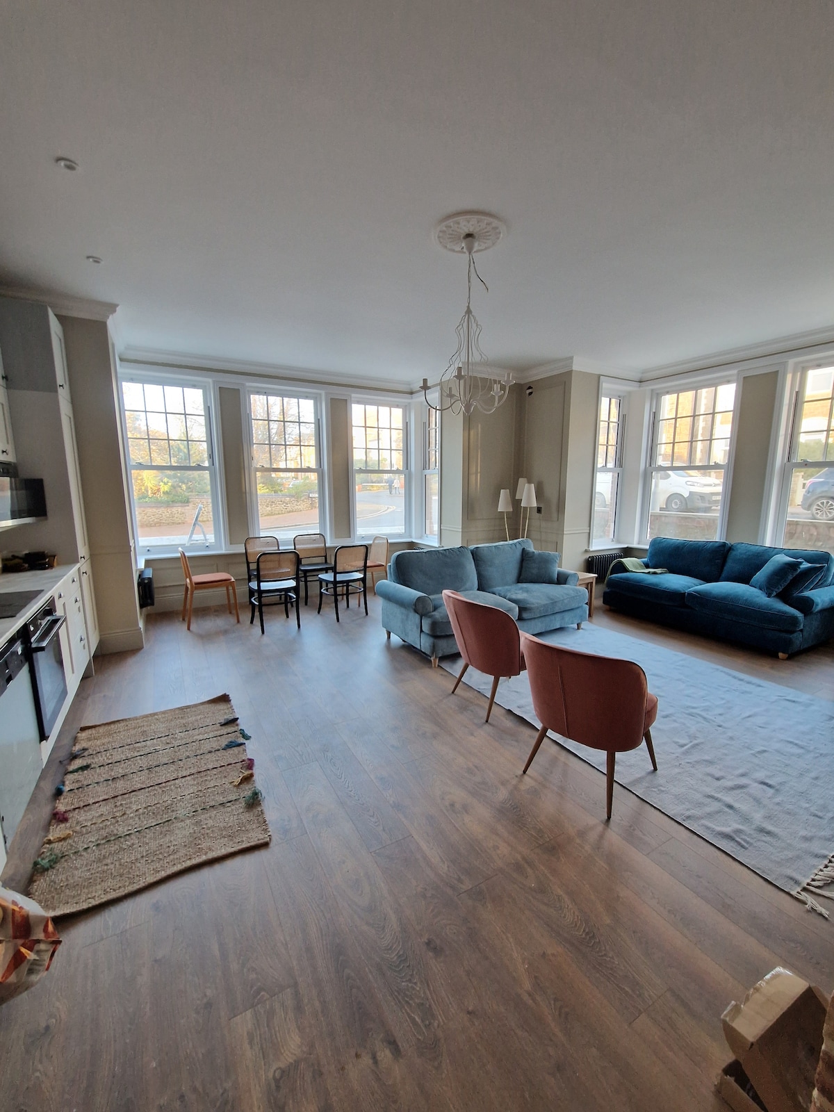 An open-plan living area features a kitchen with modern appliances, a cozy dining table with four chairs, and inviting seating options including two armchairs and a spacious sofa. Large windows allow natural light to fill the room, enhancing the warm atmosphere of the space.