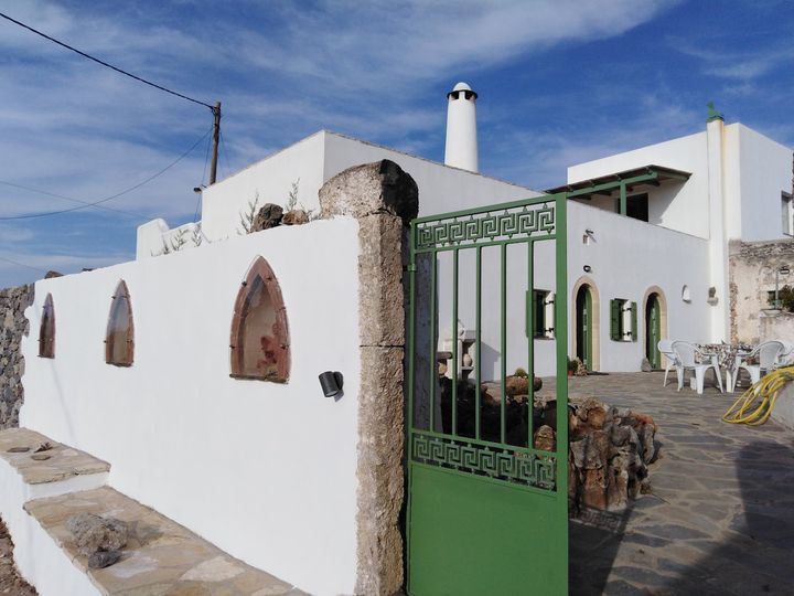 The exterior of the villa features a traditional white façade with arched windows framed in green. A wooden gate opens to a stone-paved terrace with a few outdoor chairs and a table, while a tall chimney adds height to the structure against a clear blue sky.