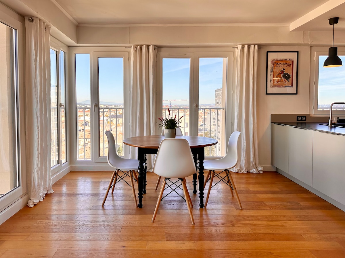 A sunlit dining area is showcased with a round wooden table surrounded by four white chairs. Large windows provide expansive city views, while light curtains frame the space. A small potted plant sits at the center of the table, adding a touch of greenery.