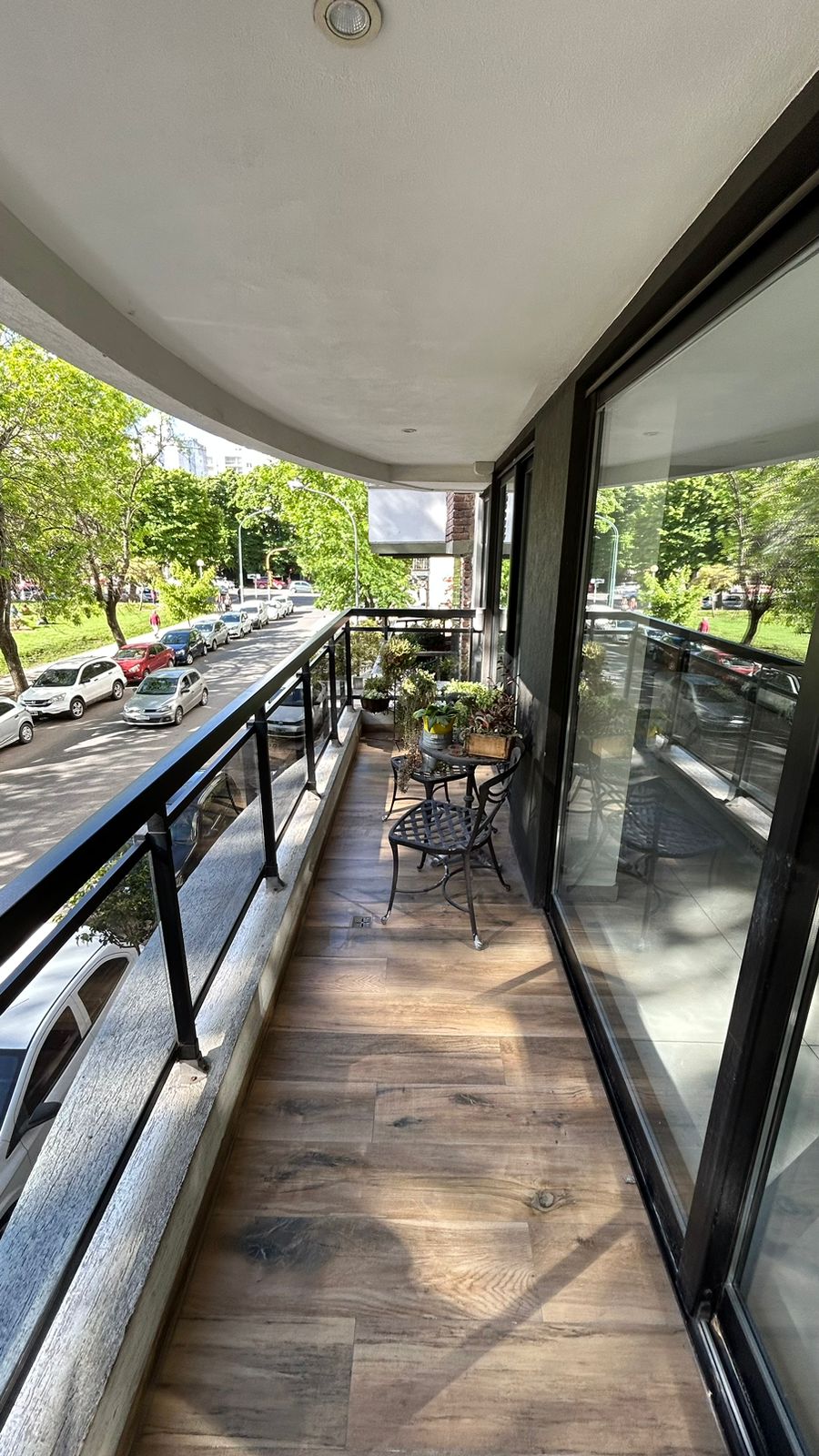 A spacious balcony is presented with wooden flooring and black railings. Two small metal chairs and a table are positioned beside potted greenery. Large glass doors offer a view of the welcoming outdoor area and surrounding tree-lined street.