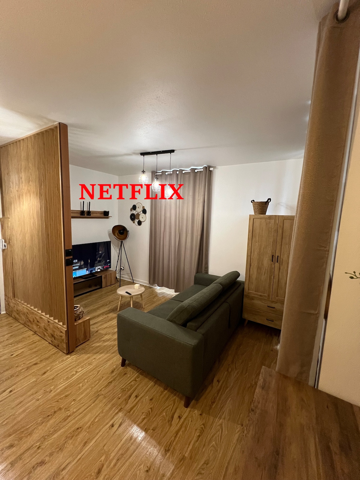 A cozy living area is presented with a green sofa positioned near a low wooden cabinet. A television is mounted on the wall, accompanied by decorative elements. Natural light filters through a floor-to-ceiling curtain, adding to the warm ambiance of the space.
