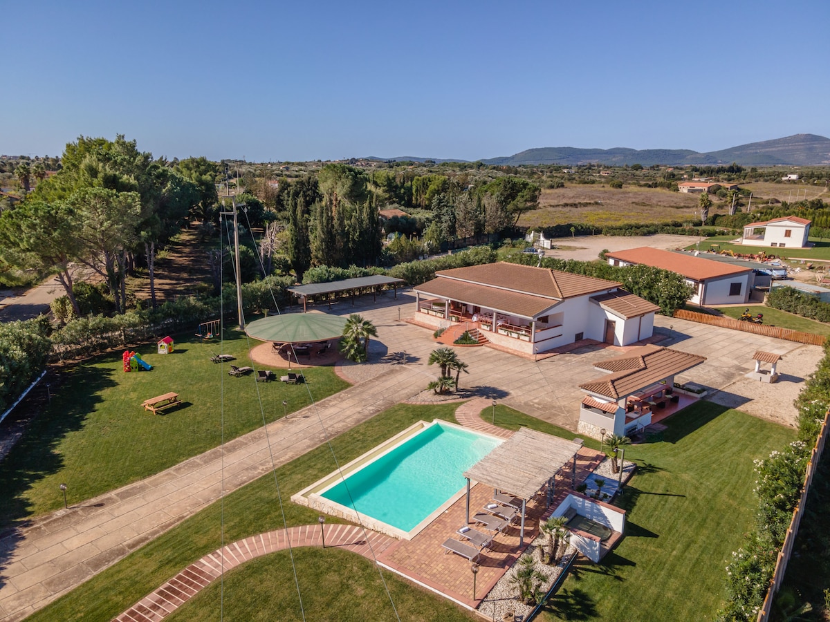 An aerial view highlights the expansive outdoor area, featuring a swimming pool surrounded by greenery. Shaded seating areas and a gazebo invite relaxation, while a well-maintained lawn is interspersed with picnic spots. The villa's structure can be seen in the background, with nearby mountains framing the scene.