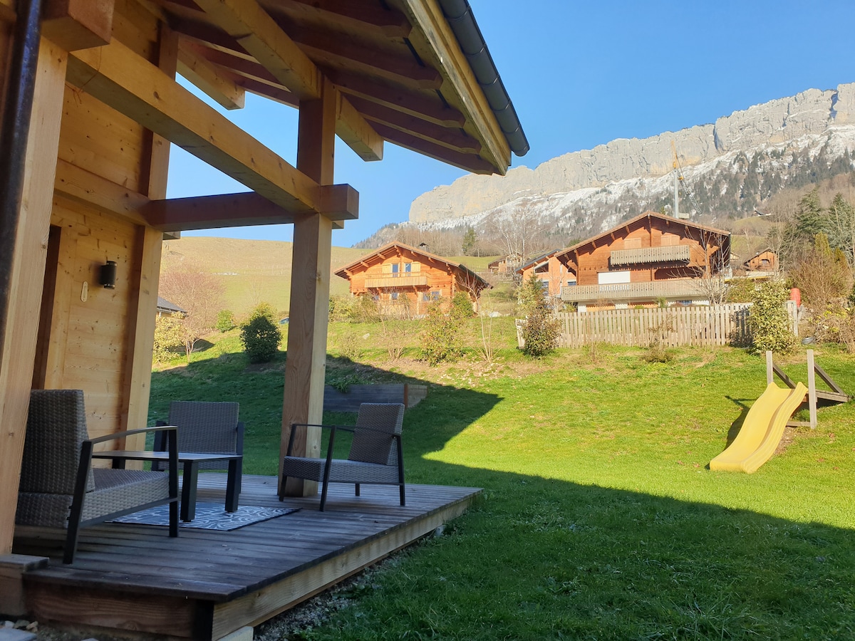 A wooden deck area features two chairs and a small table, inviting outdoor relaxation. A grassy space extends toward a gently sloped slide, with scenic mountain views in the background. Nearby chalets are visible among the lush greenery, creating a serene and natural environment.