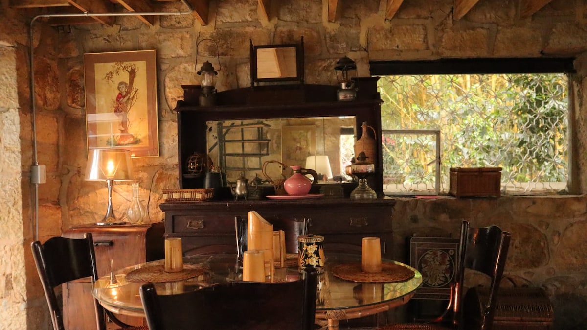 A rustic dining area features a round table set with woven placemats. An antique sideboard holds decorative items and candles, while a window offers a glimpse of the lush greenery outside. Warm lighting creates a cozy ambiance throughout the stone-walled space.