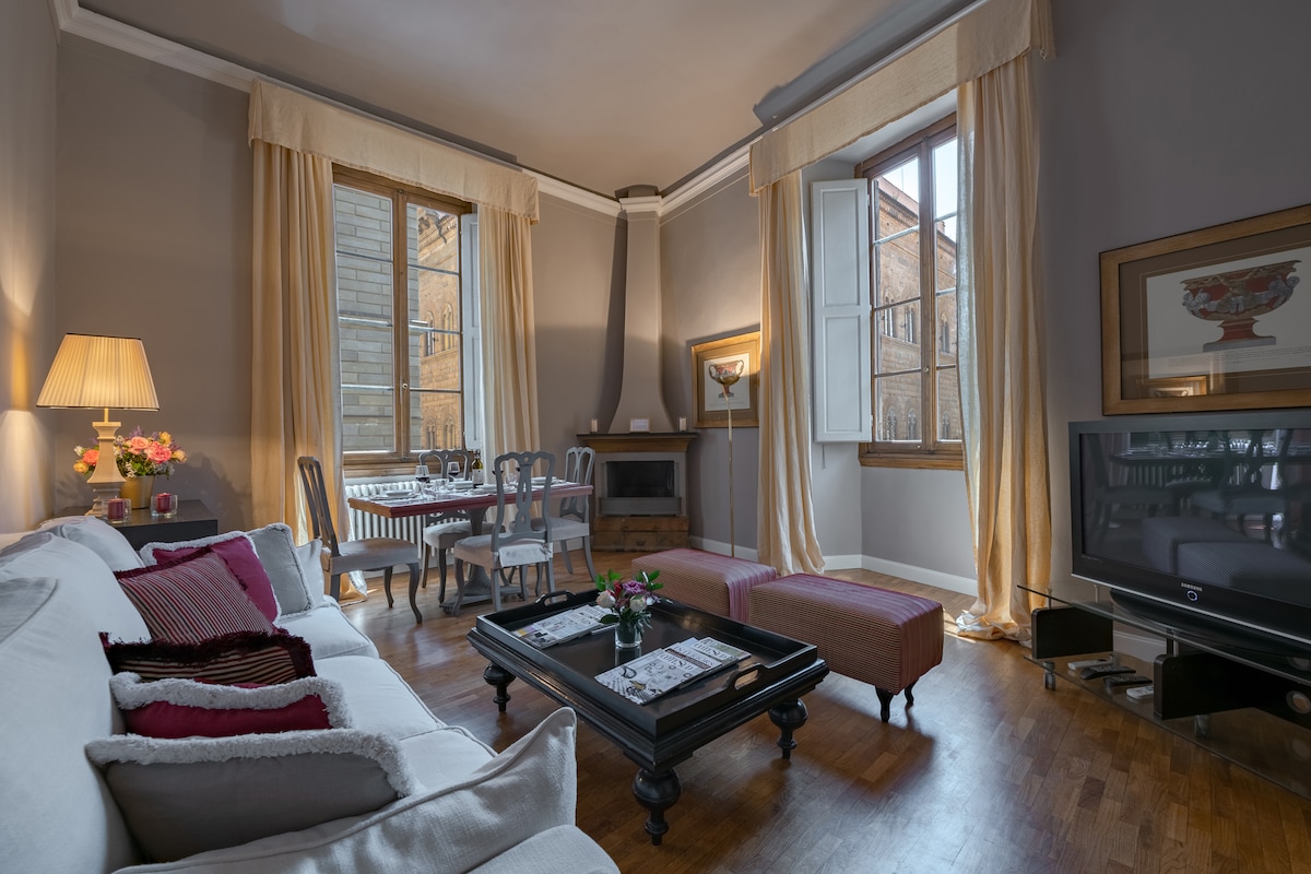 A spacious living area is illuminated by natural light from large windows adorned with soft curtains. A comfortable sofa, accented with cushions, complements a low coffee table. A dining table for four is positioned nearby, creating an inviting space for gatherings.