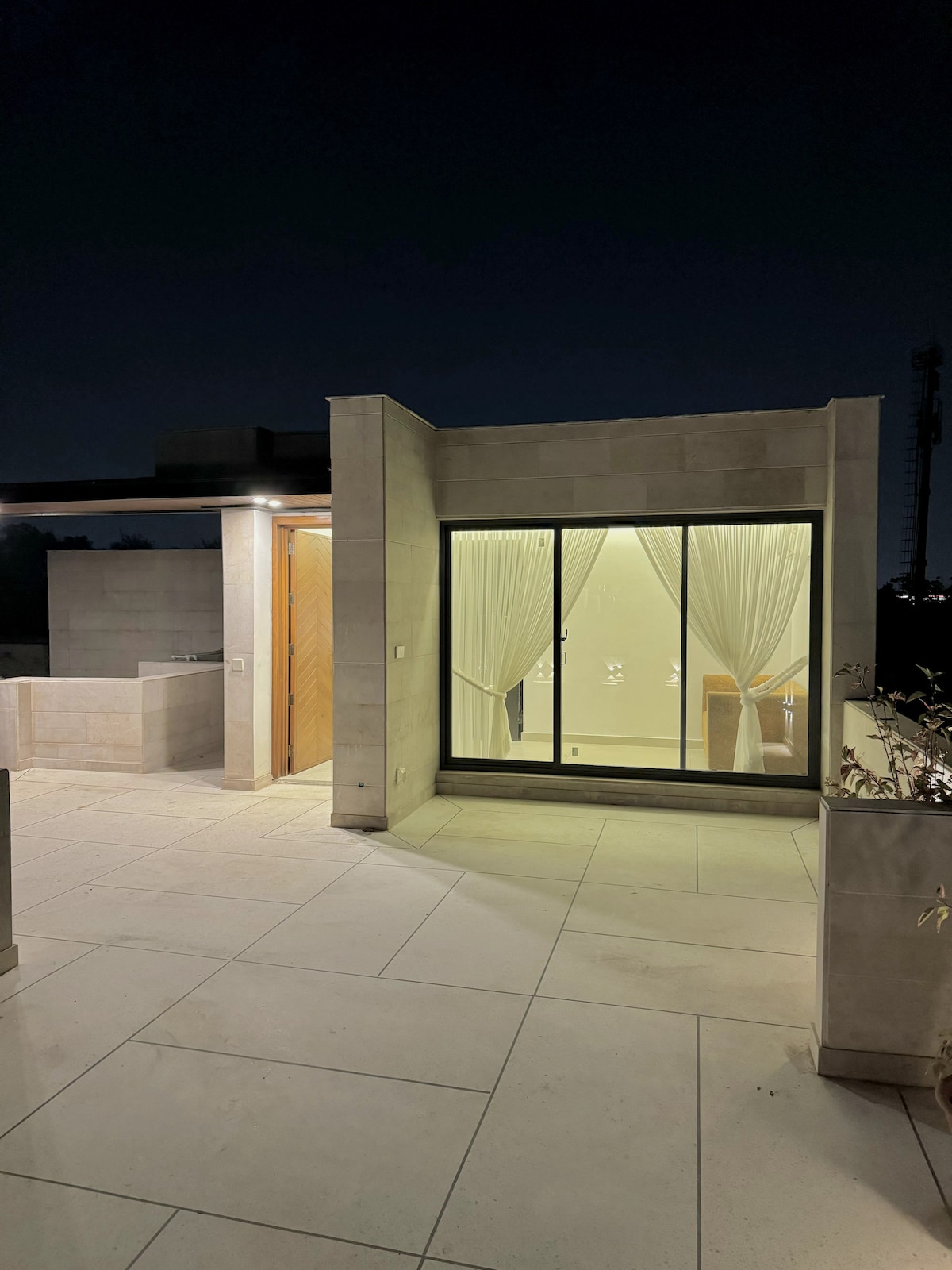 An outdoor area is illuminated against the night sky, showcasing modern architecture. Large glass doors provide a view into the interior space, which is softly lit with sheer curtains framing the window. Clean lines and a light-colored stone floor contribute to the contemporary aesthetic.