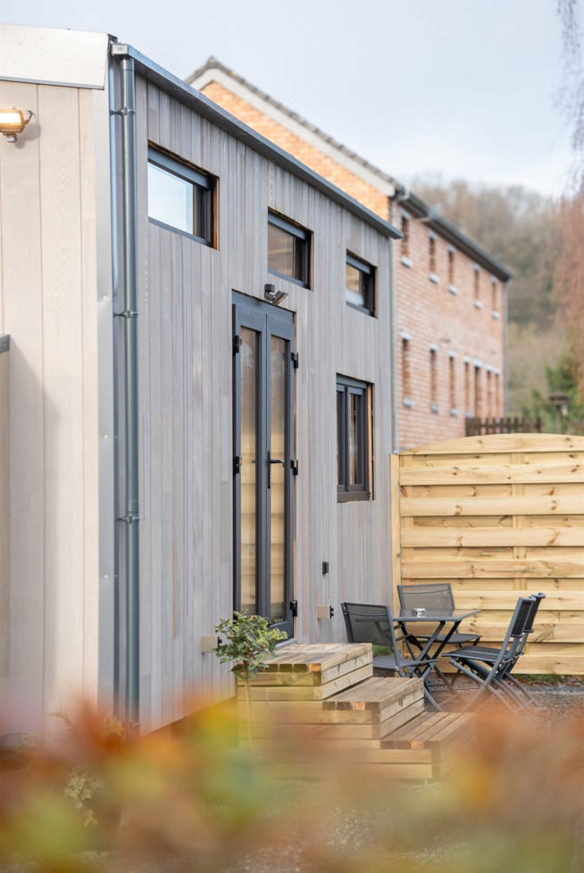 The exterior of the tiny house features wooden cladding and large windows, allowing natural light to enter. A small outdoor seating area with black chairs is positioned beside the entrance, surrounded by landscaped foliage. The tranquil atmosphere is accentuated by the nearby natural surroundings.