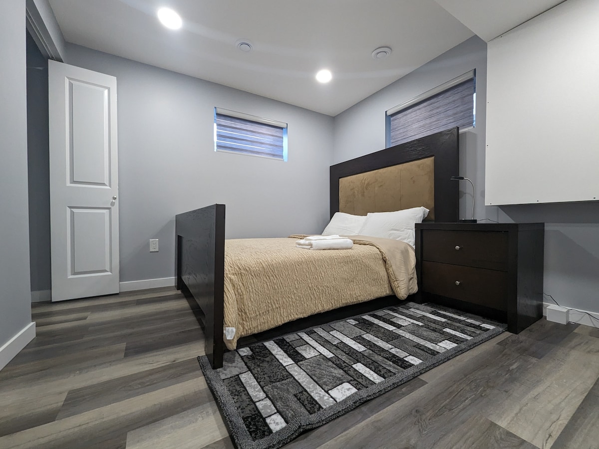 A spacious bedroom features a comfortable bed with a warm-toned blanket. Soft lighting illuminates the room, highlighting the neutral wall colors and wood flooring. A bedside table and a textured area rug add to the room's comfort, while windows with light-filtering blinds provide natural light.