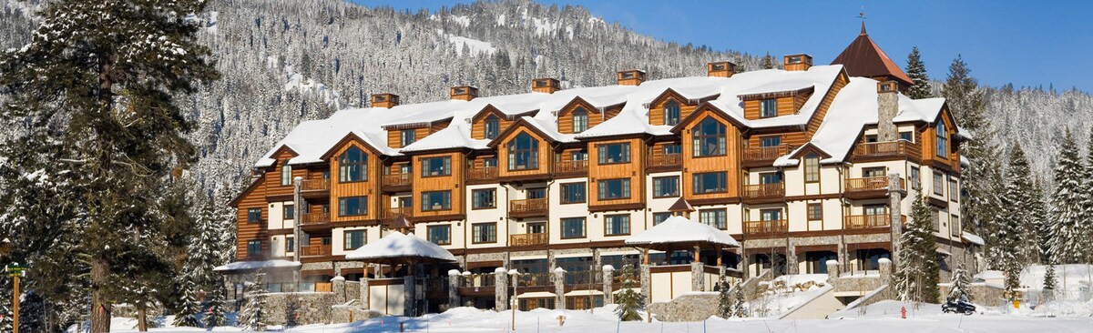 A multi-story building is set against a backdrop of snow-covered mountains. The structure features wooden accents and large windows, reflecting a warm and inviting aesthetic. Snow blankets the ground, enhancing the winter atmosphere of the resort.