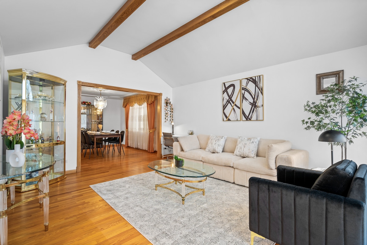A living area showcases an elegant, light-colored sofa paired with a black armchair. A glass coffee table sits atop a patterned rug, while wooden beams accent the ceiling. A dining space can be seen through an archway, complementing the overall spacious feel.