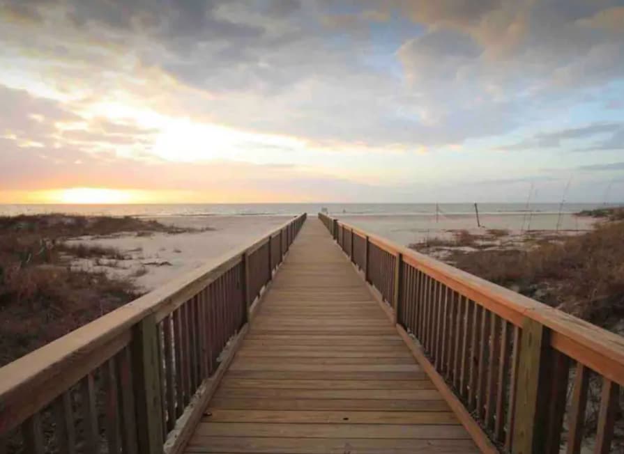 A wooden boardwalk extends toward the ocean, flanked by sandy beach and coastal vegetation. The horizon displays soft hues of orange and purple as the sun sets, creating a serene pathway leading to the water's edge.