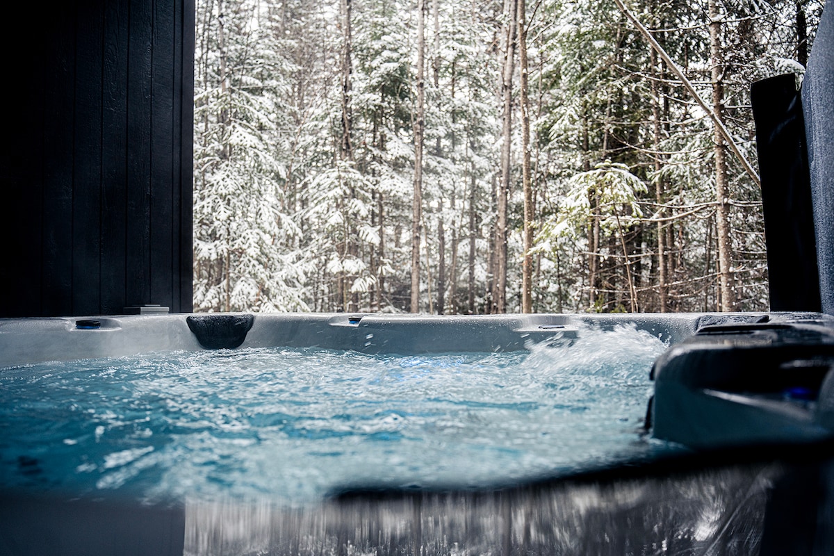 A hot tub is visible, with bubbling water reflecting the light. Surrounding it, snow-covered trees create a serene backdrop, hinting at a peaceful outdoor environment. The image captures the tranquility and natural beauty of the setting.