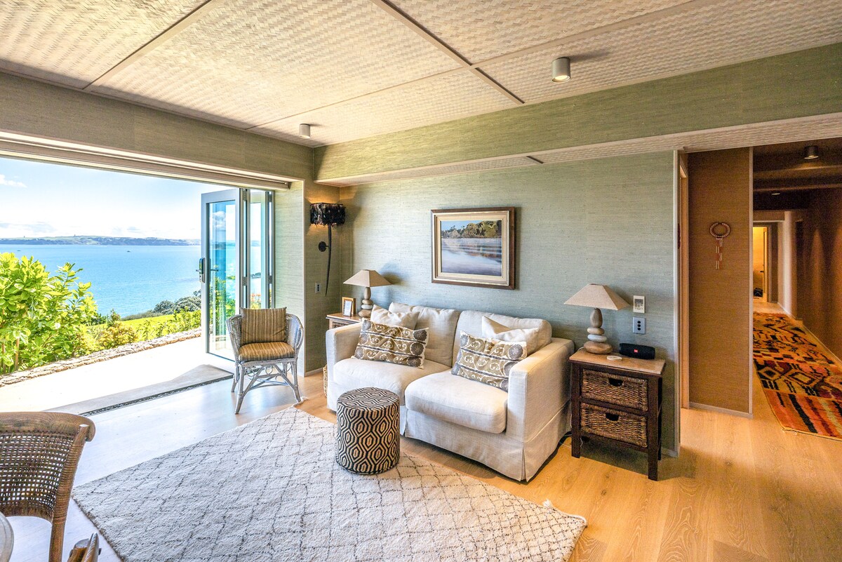 A living area features a light-colored sofa with decorative cushions, complemented by a circular ottoman. Natural light fills the space from large glass doors that open to scenic views of the sea. A framed artwork adorns the wall, adding a touch of elegance.