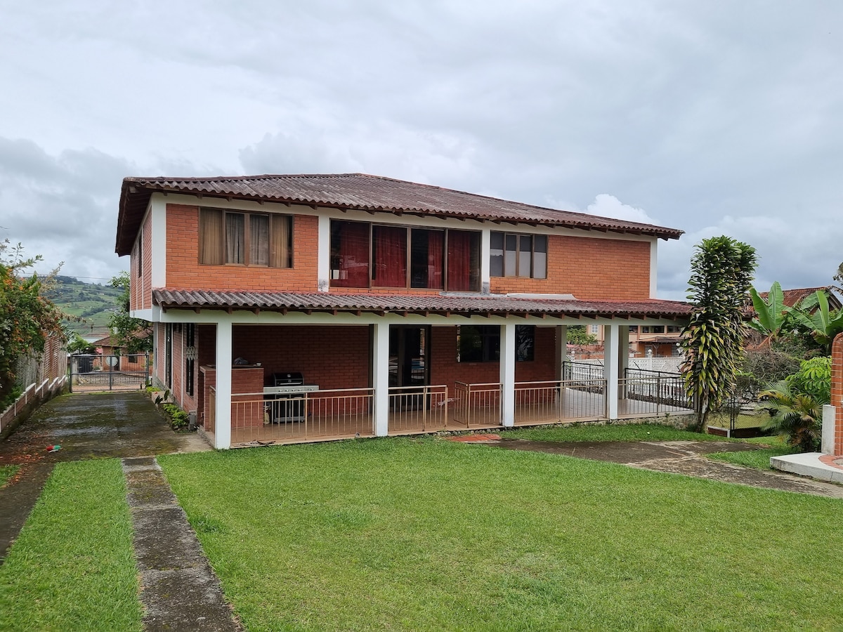 A two-story brick house is shown, featuring a covered porch area with seating. Large windows provide natural light to the interior. Surrounding green lawns and pathways lead through a landscaped yard, with a backdrop of hills and cloudy skies.