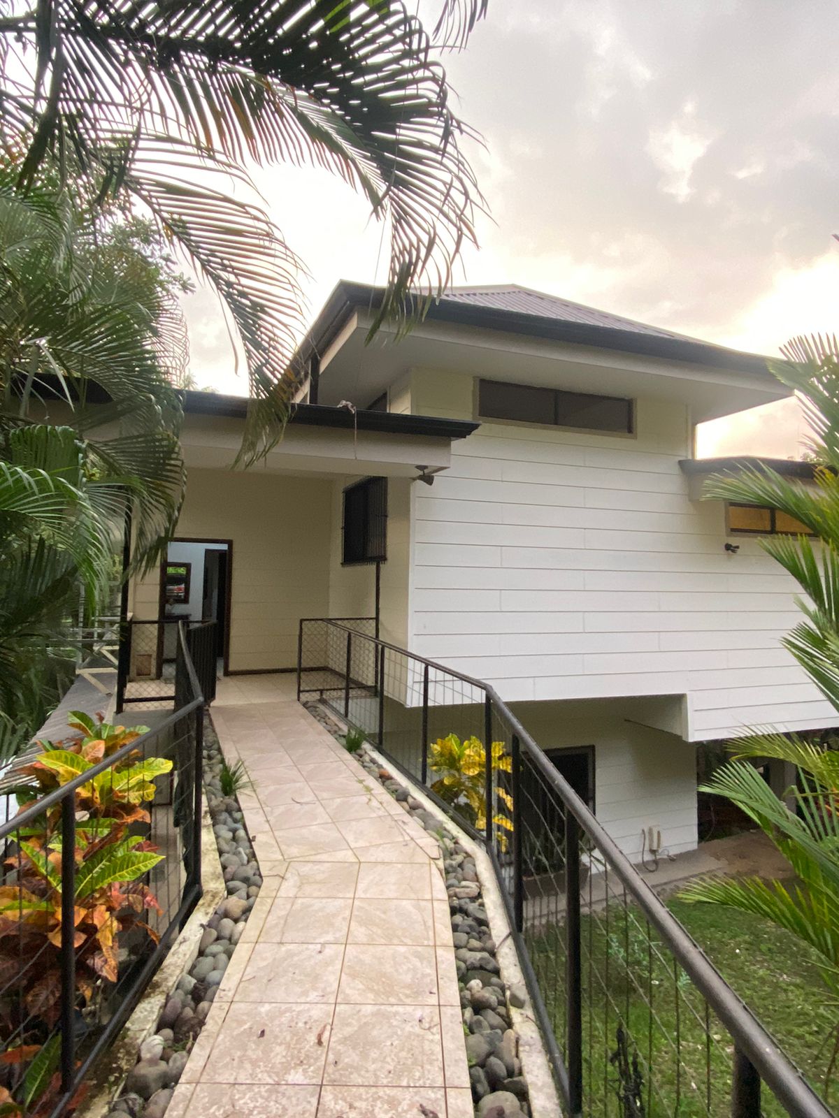 A paved pathway lined with tropical plants leads to the entrance of the house. The exterior features a combination of white and dark materials, with large windows providing ample natural light. The structure is surrounded by lush greenery.
