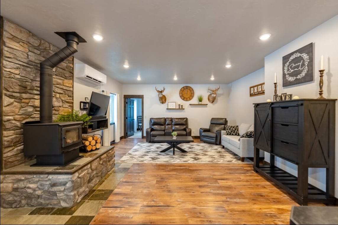 A spacious living area features a stone fireplace with a wood stove and stacked logs beside it. Comfortable seating includes a dark leather couch and a recliner, complemented by a patterned rug. Wall decor includes mounted antlers and tasteful artwork, enhancing the cozy ambiance.