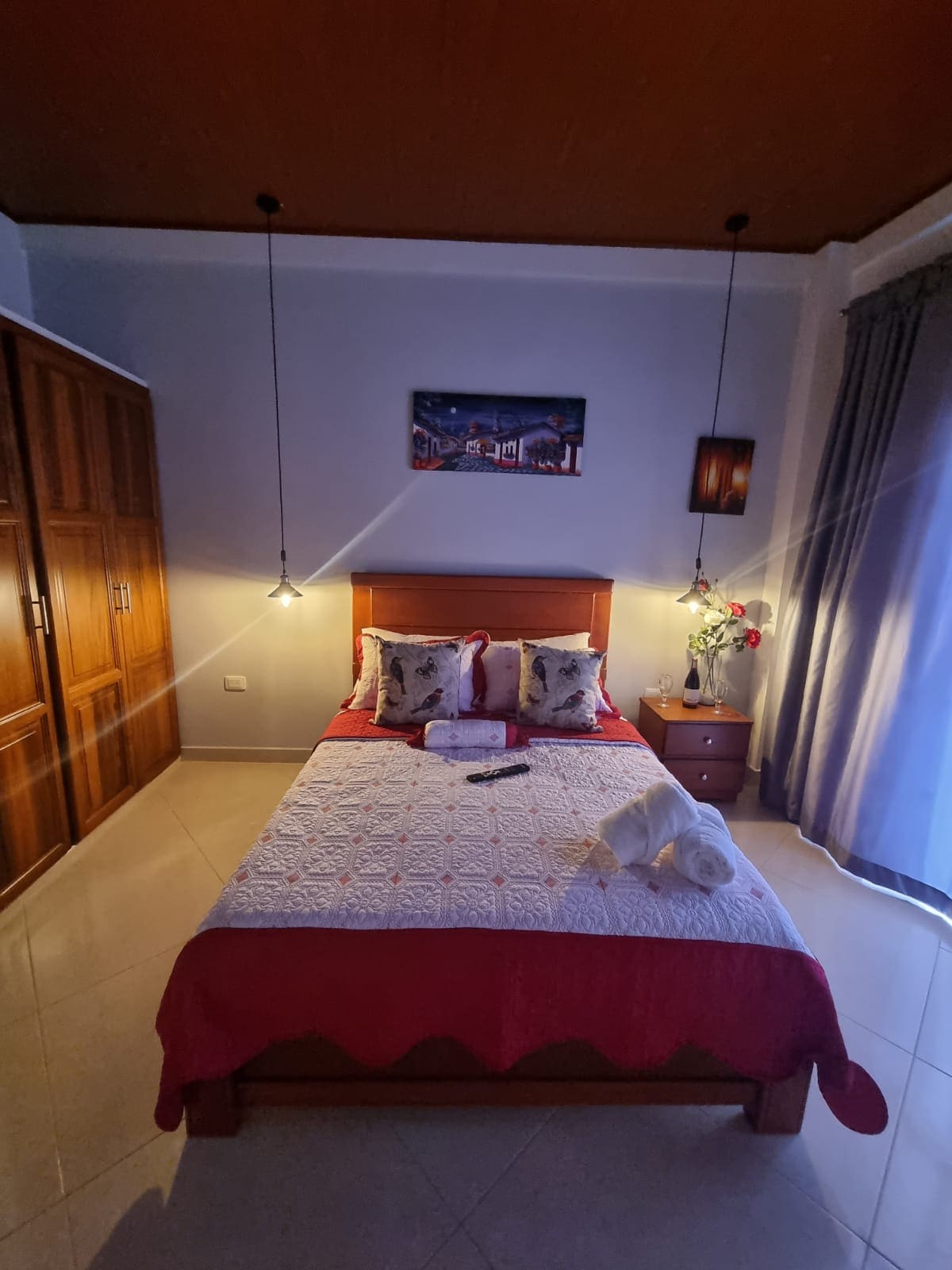 A cozy bedroom is presented with a spacious bed dressed in white and red linens. Two pendant lights illuminate the room, highlighting decorative pillows. A wooden wardrobe stands against the wall, and a window draped with gray curtains allows natural light to enter.