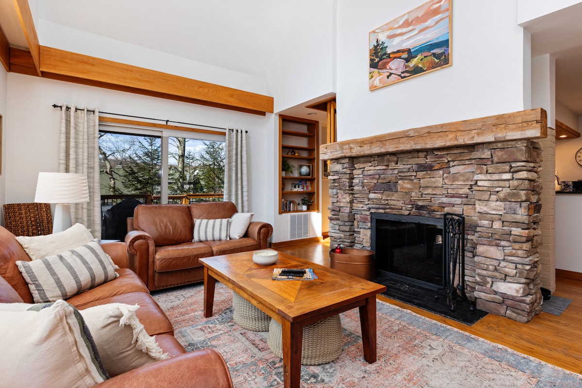 The living room features a stone fireplace as a central element, surrounded by comfortable leather seating. A wooden coffee table sits in front, and large windows allow natural light to fill the space, offering a view of the outdoors. Artwork adorns the wall above the fireplace.