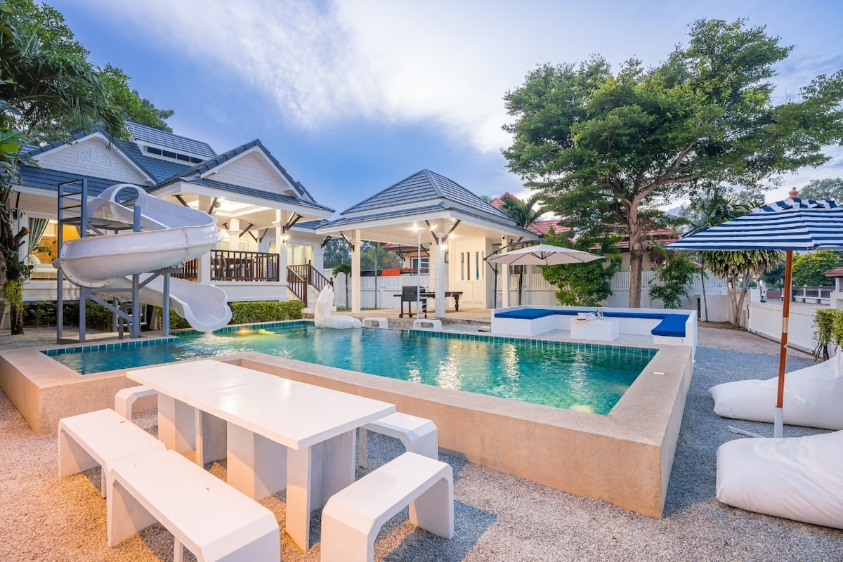 An inviting outdoor area features a large swimming pool with an integrated slide. A spacious dining table is located nearby, surrounded by comfortable seating. Sun umbrellas provide shade, while lush trees frame the scene, creating a relaxed setting for outdoor gatherings.