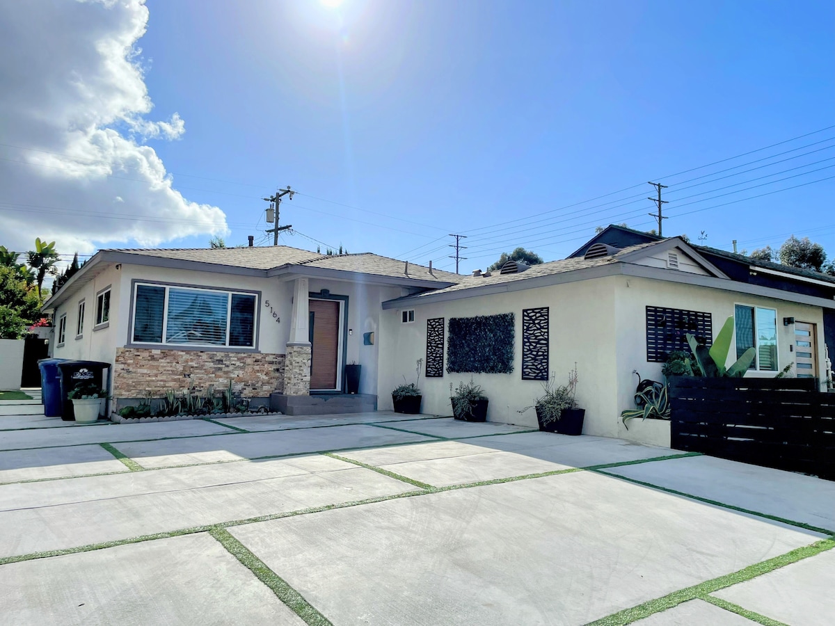 A single-story home is presented, featuring a light-colored exterior with stone accents. The front porch is visible, along with large windows that allow natural light inside. A spacious driveway is shown, lined with green grass strips, providing ample parking space.