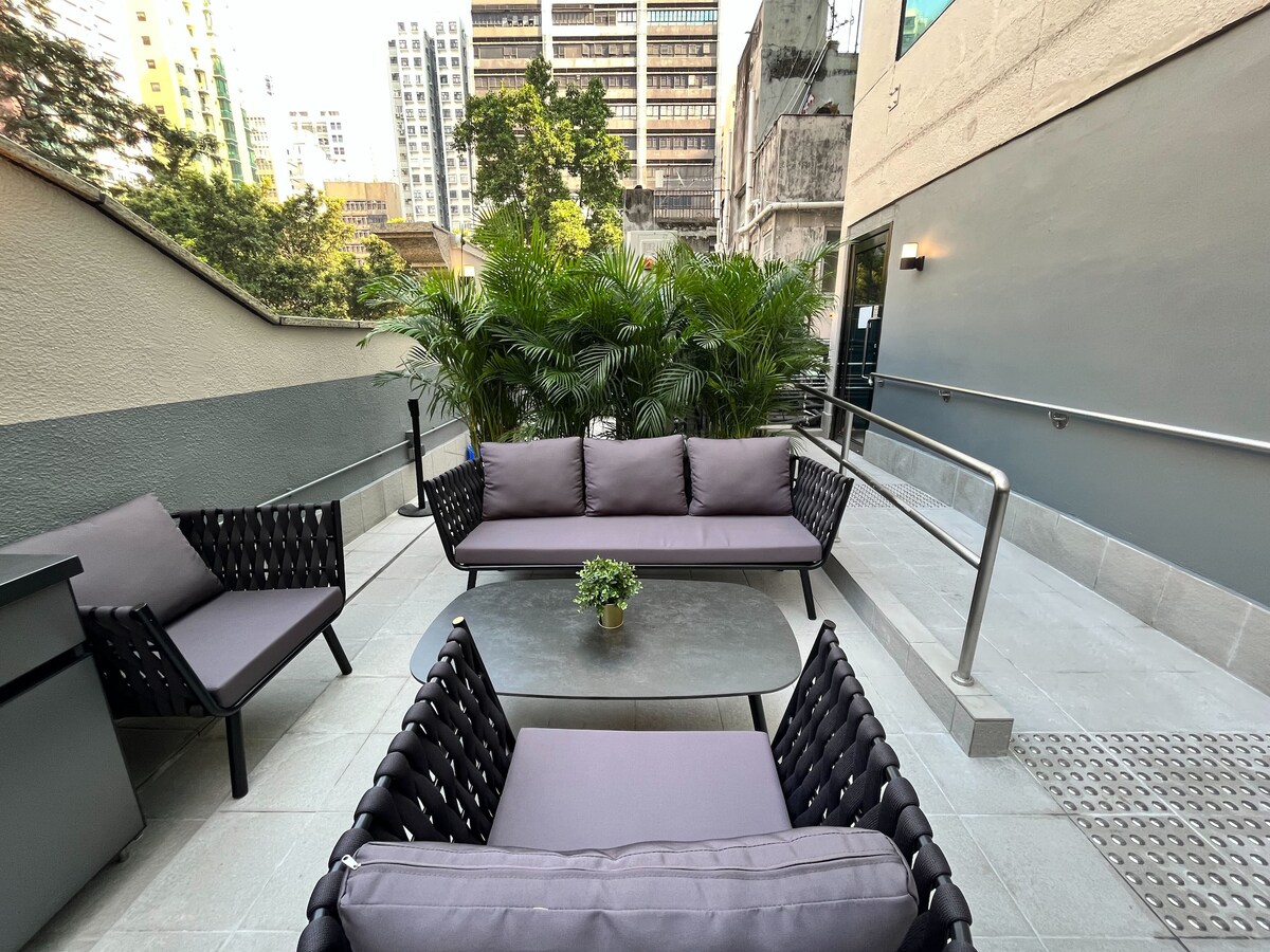 A modern outdoor seating area is presented with two armchairs and a sofa, all featuring soft gray cushions. A small round table sits in the center. Lush greenery surrounds the space, providing a refreshing and tranquil atmosphere in an urban setting.