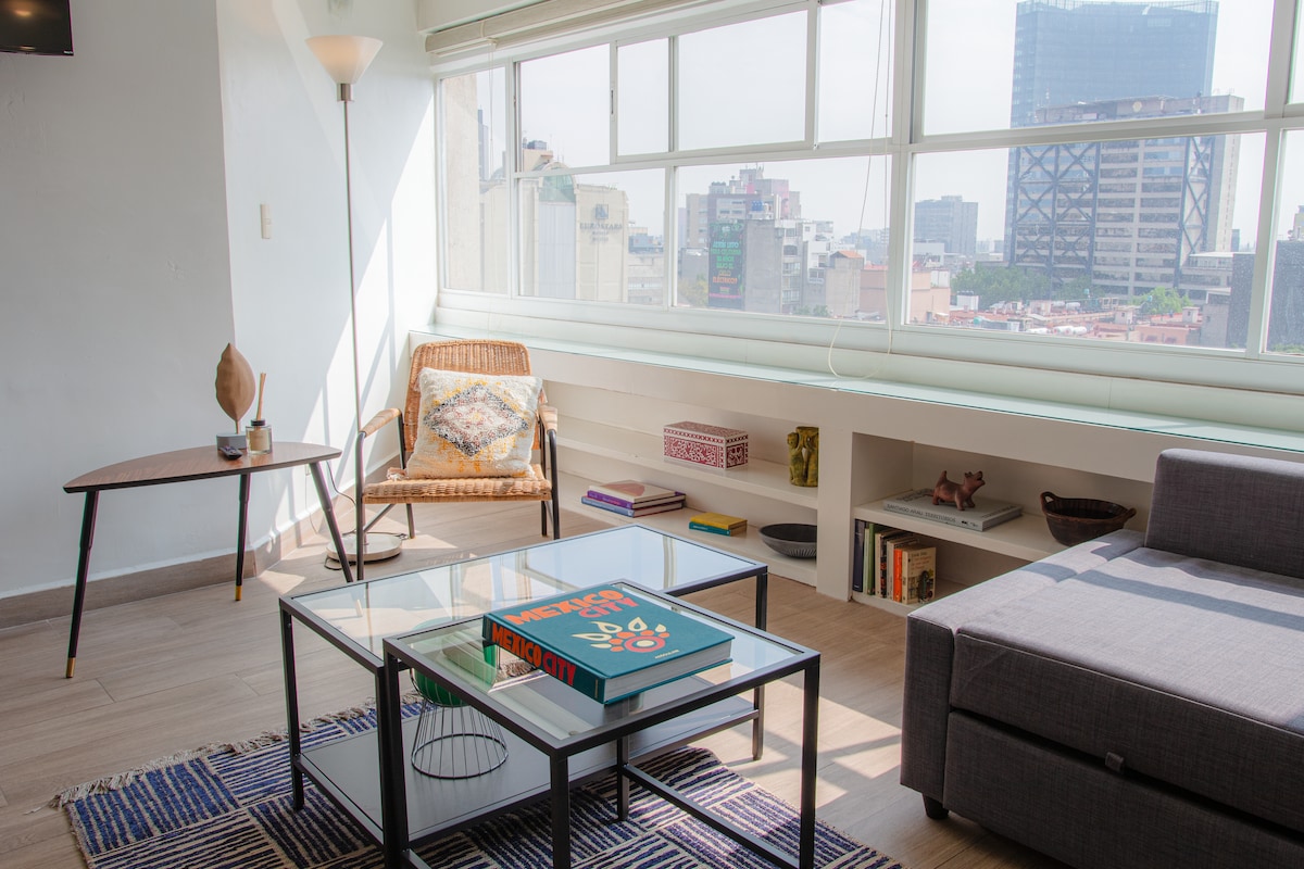 A bright living area features large windows that provide a panoramic view of the city skyline. A minimalist coffee table sits atop a striped area rug, complemented by a comfortable sofa and a stylish chair. Shelves with decorative items are neatly arranged against the wall.