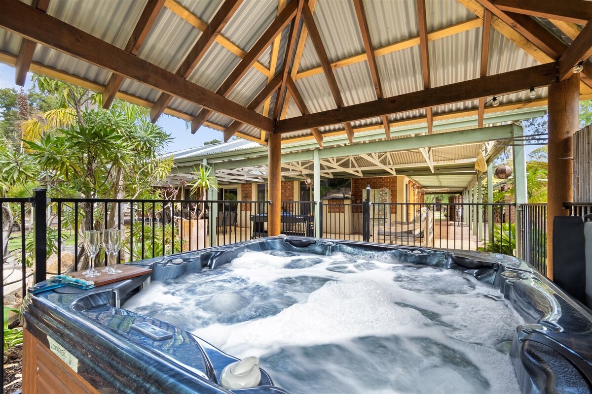 A covered al fresco area features a six-seat hot tub surrounded by a wooden deck and railings. Lush greenery and palm trees enhance the outdoor ambiance, while a shaded roof provides protection from the elements.