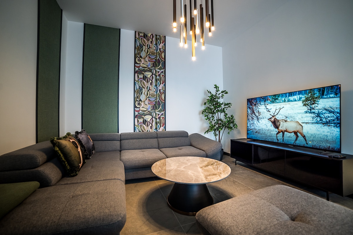 A modern living room is presented with a gray sectional sofa arranged around a round coffee table. A large television displays a serene outdoor scene. Green accents are provided by a potted plant and wall decorations with natural motifs, while multiple light fixtures hang elegantly from the ceiling.