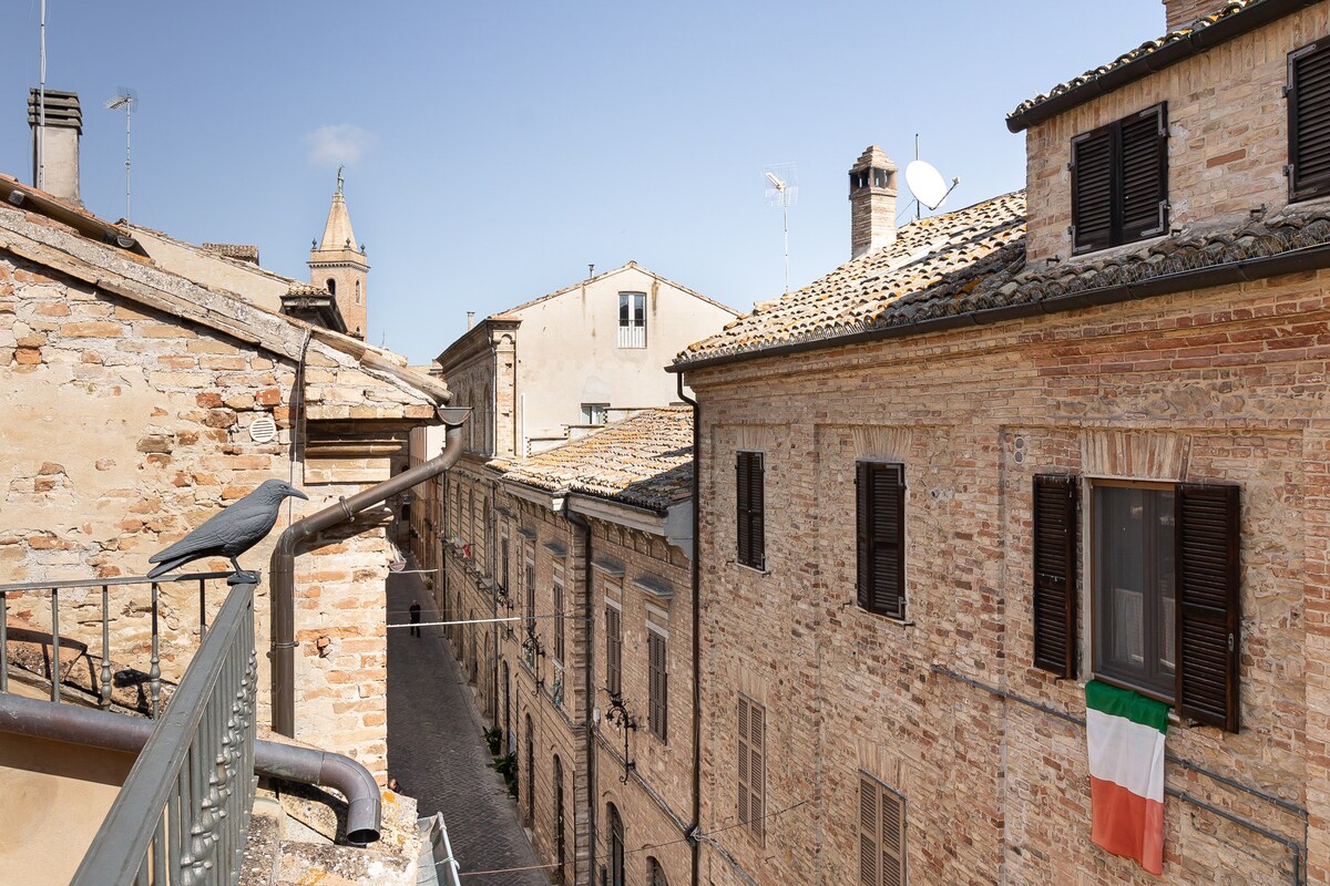 A view from a balcony shows a charming street lined with historic buildings featuring natural stone facades. A flag of Italy hangs from one of the windows. The scene is complemented by a clear blue sky and distant rooftops, enhancing the village atmosphere.