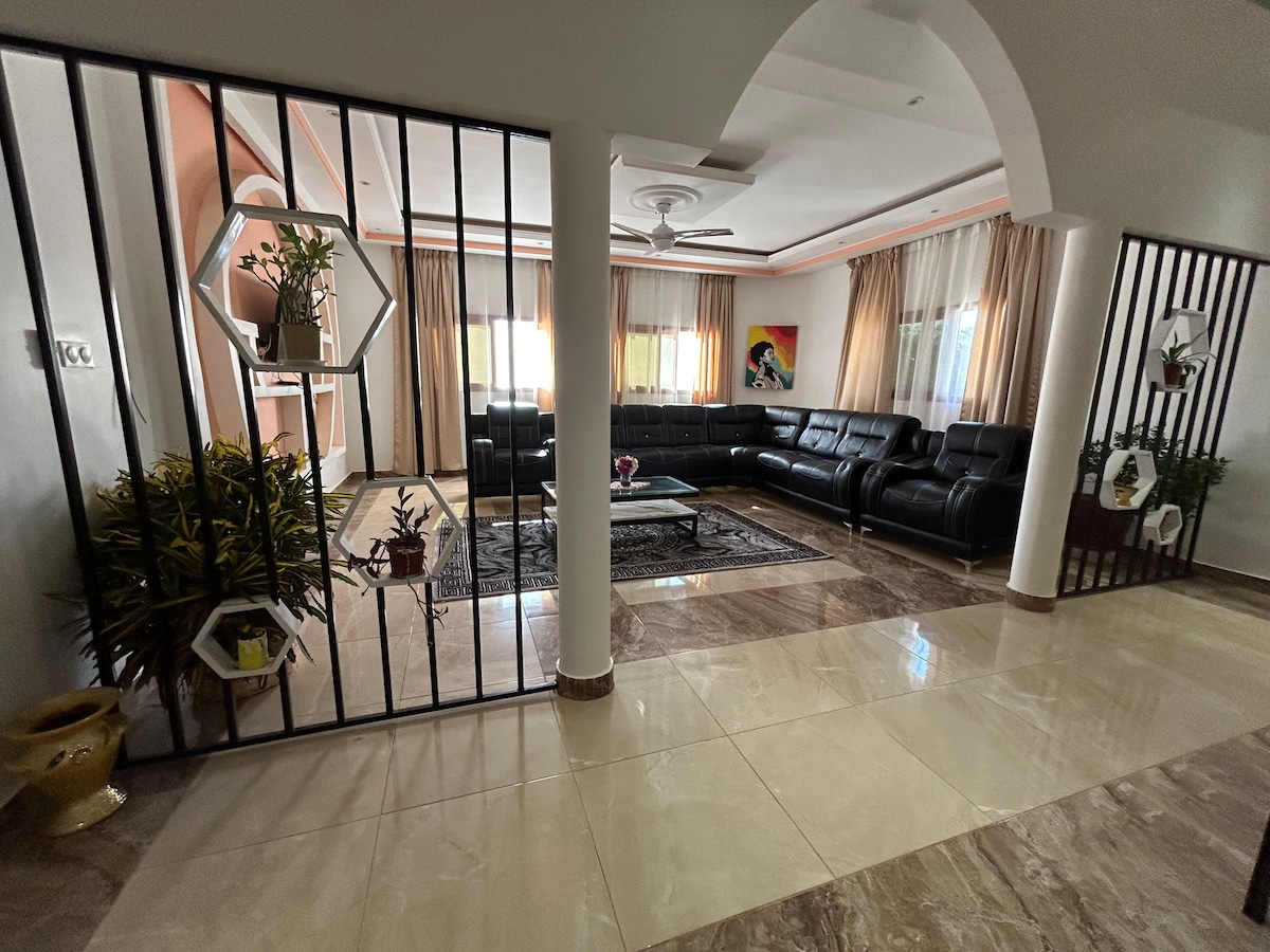 A spacious living area features a black sectional sofa arranged around a central glass coffee table. Light-colored curtains frame the windows, allowing natural light to fill the room. Decorative plants are positioned near the entrance, enhancing the welcoming ambiance of the space.