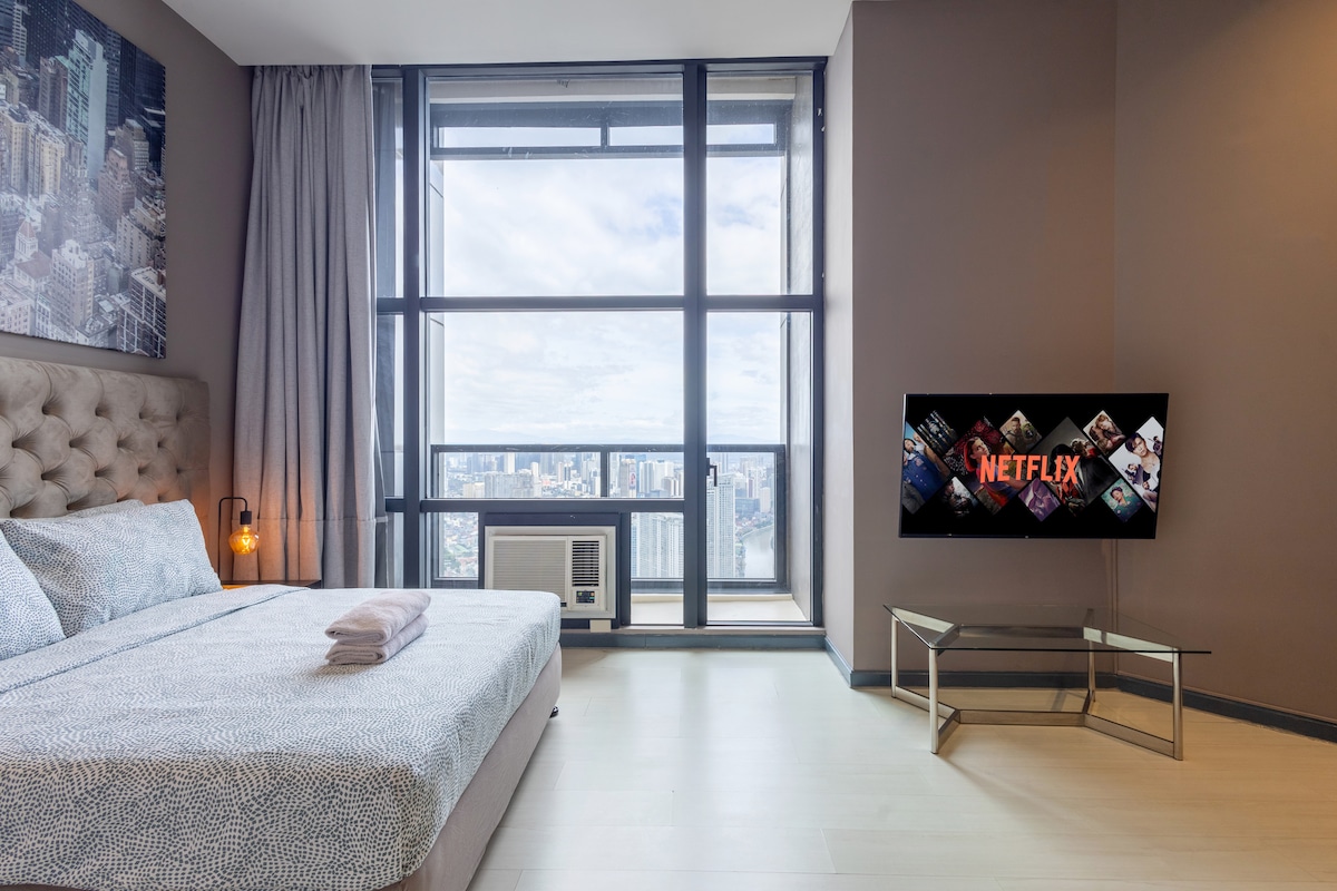 A spacious studio apartment is highlighted by a large window offering views of the city skyline. A comfortable bed with a plush headboard is positioned beside the window, and a 65" TV displaying Netflix is mounted on the wall across from the bed.