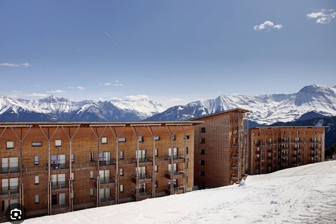 A wooden building structure is surrounded by snow-covered terrain and majestic mountain views. Large windows characterize the facade, allowing for ample natural light. The landscape displays a mix of snow and mountains under a clear blue sky, providing a serene and tranquil atmosphere.