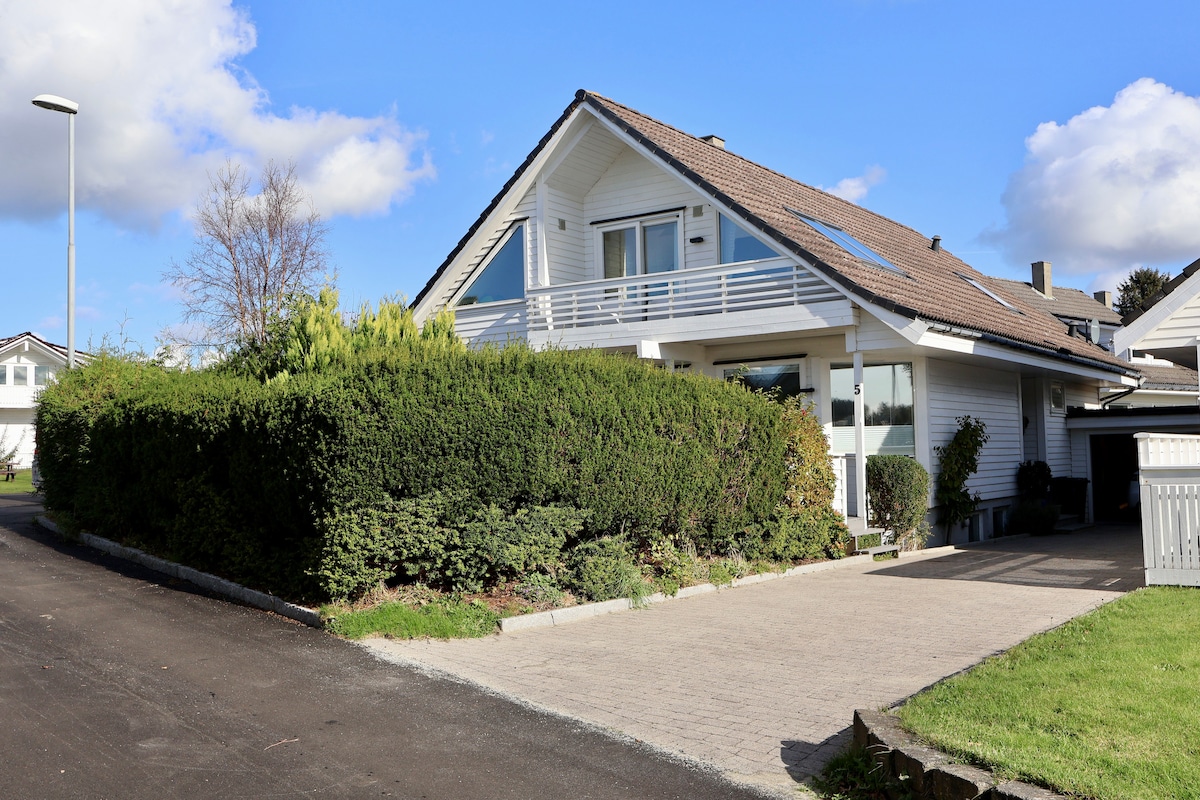 A spacious white house is set against a clear blue sky, surrounded by well-maintained greenery. A paved driveway leads up to the entrance, which features large windows and a balcony. The landscaping includes neatly trimmed hedges and shrubs, contributing to a calm environment.