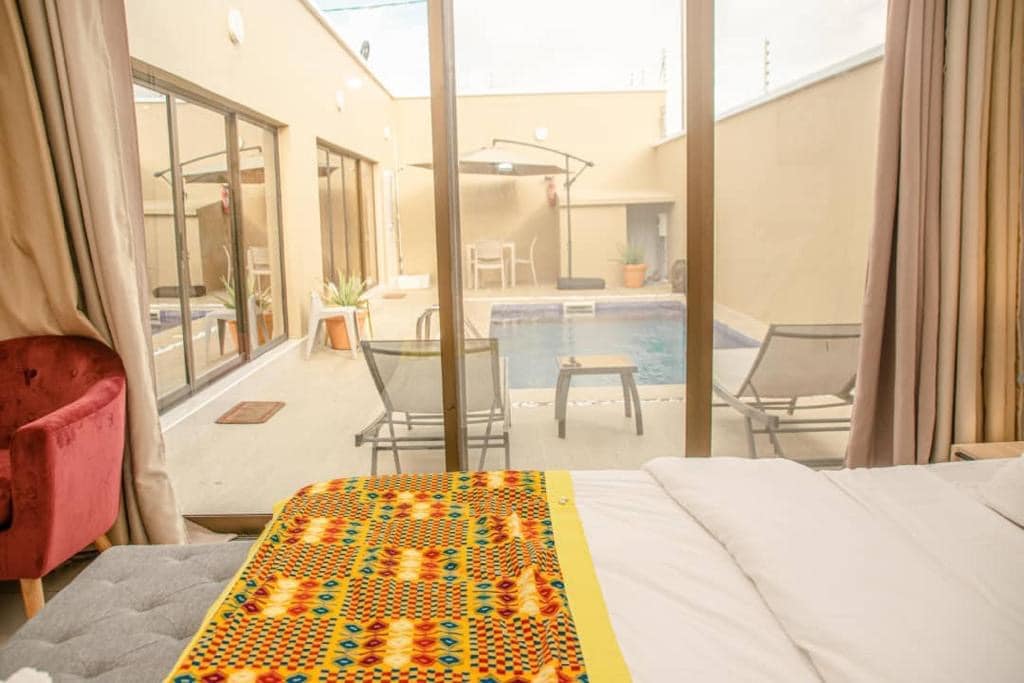 A bedroom features a spacious bed dressed in white linens, with a colorful patterned blanket. Large glass doors offer a view of a private patio with a pool, lounge chairs, and plants, allowing natural light to fill the room.