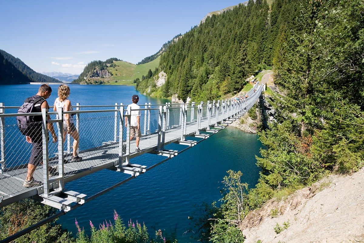 A suspension bridge spans a tranquil lake, surrounded by lush green mountains and pine trees. Three individuals walk across the bridge, with a clear blue sky above. The landscape reflects natural serenity, inviting exploration of the scenic environment.