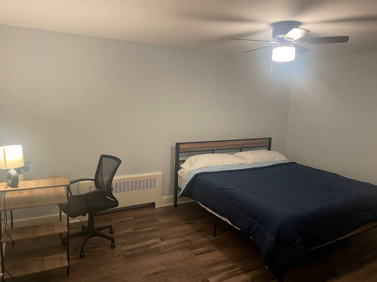 A spacious bedroom features a king-size memory foam bed with white linens, complemented by a dark blue blanket. A desk with an ergonomic chair is positioned nearby, illuminated by a table lamp. The room is equipped with a ceiling fan and has warm wood flooring.