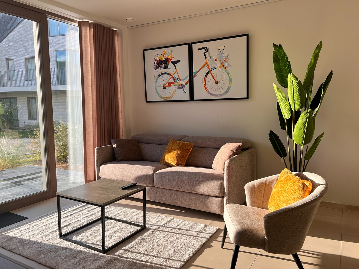 A welcoming living area is furnished with a light brown sofa and a cream-colored armchair. A coffee table occupies the center, and colorful artwork featuring a bicycle adorns the wall. Natural light floods the space through large glass doors, complemented by indoor plants nearby.