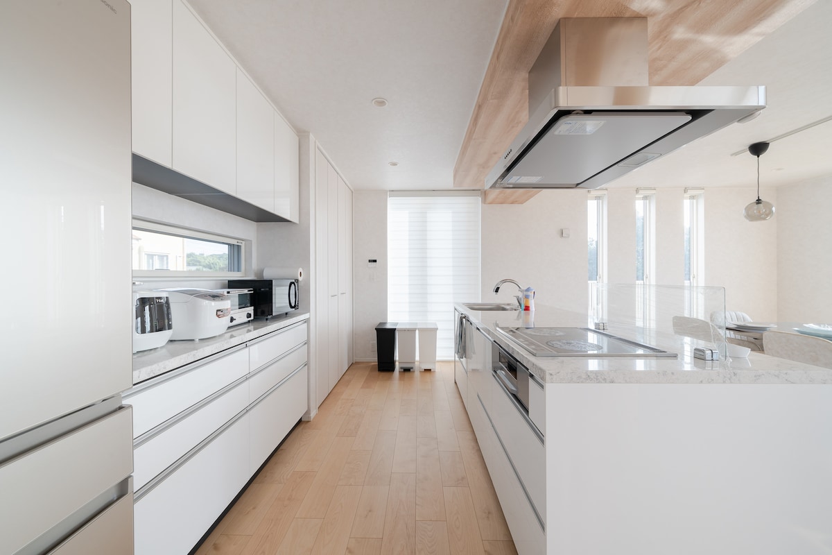 A modern kitchen is presented with sleek white cabinetry and a spacious countertop. Stainless steel appliances, including a refrigerator and microwave, are visible. Natural light filters through the windows, enhancing the clean lines and minimalist design of the space.