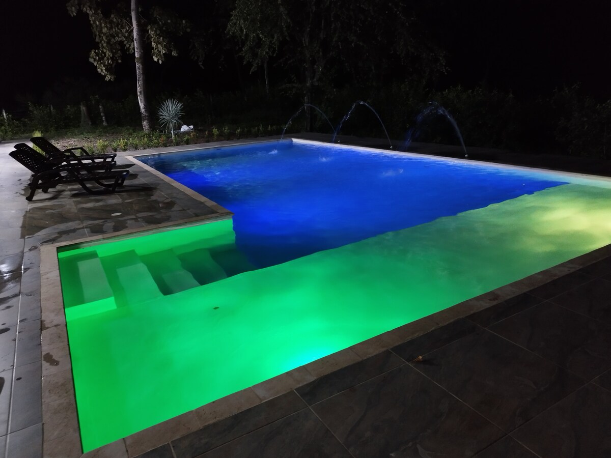 The outdoor pool features illuminated water in shades of blue and green, enhancing the evening ambiance. Two lounge chairs sit alongside the pool, providing a space for relaxation. Water features add movement, creating a calm and serene outdoor environment.