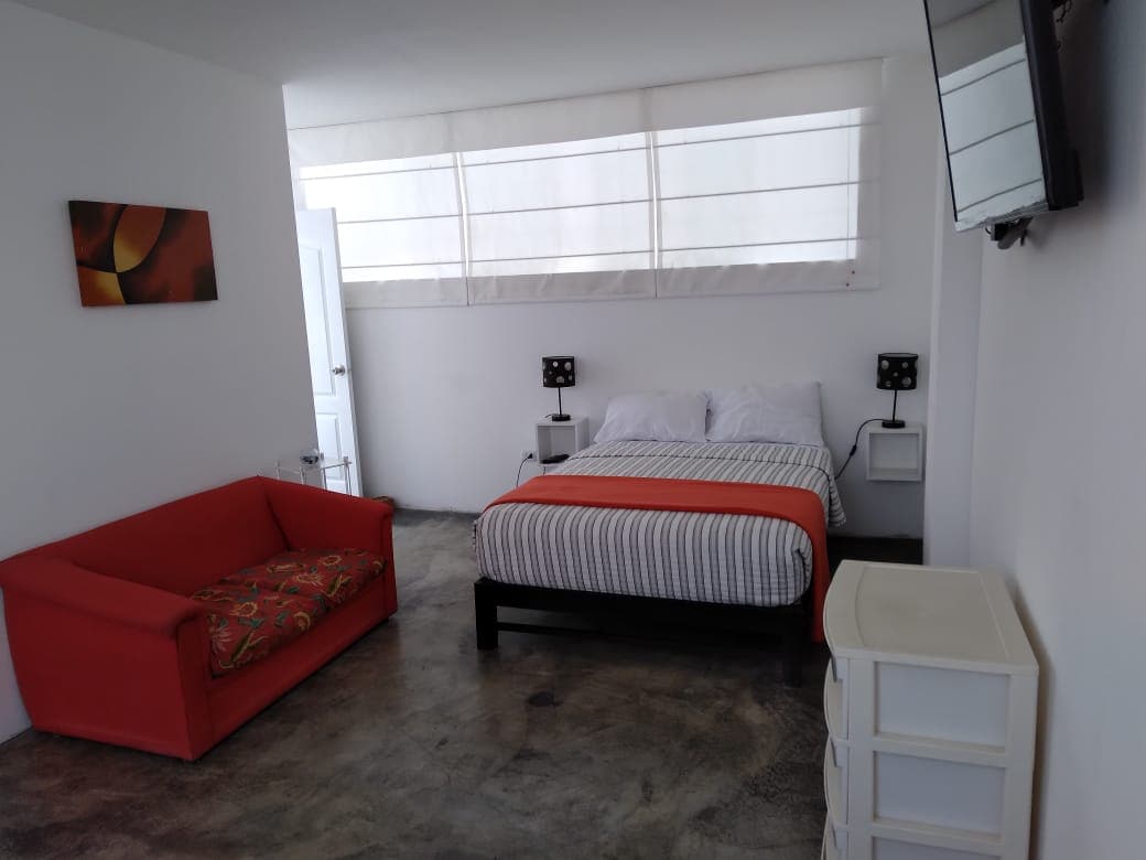 A bright room features a spacious bed with striped linens, flanked by nightstands and stylish lamps. A vibrant red sofa adds a pop of color, while large windows allow natural light to fill the space. A dresser is positioned against the wall, providing additional storage.