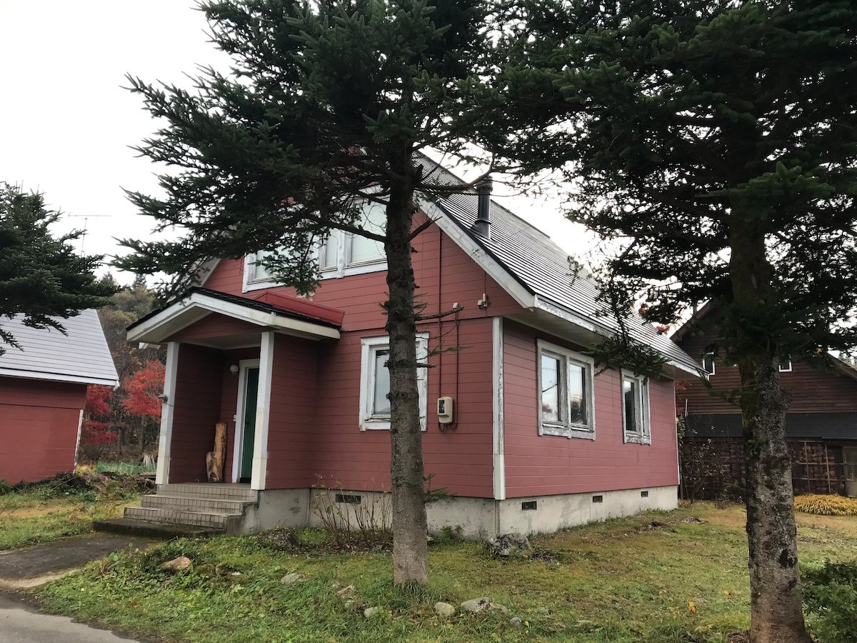 A two-story house with a pink exterior and a sloped gray roof is framed by trees in the foreground. Large windows offer views of the surroundings, while a front porch with a single step leads to the entrance. The grassy area around the house is well-kept.