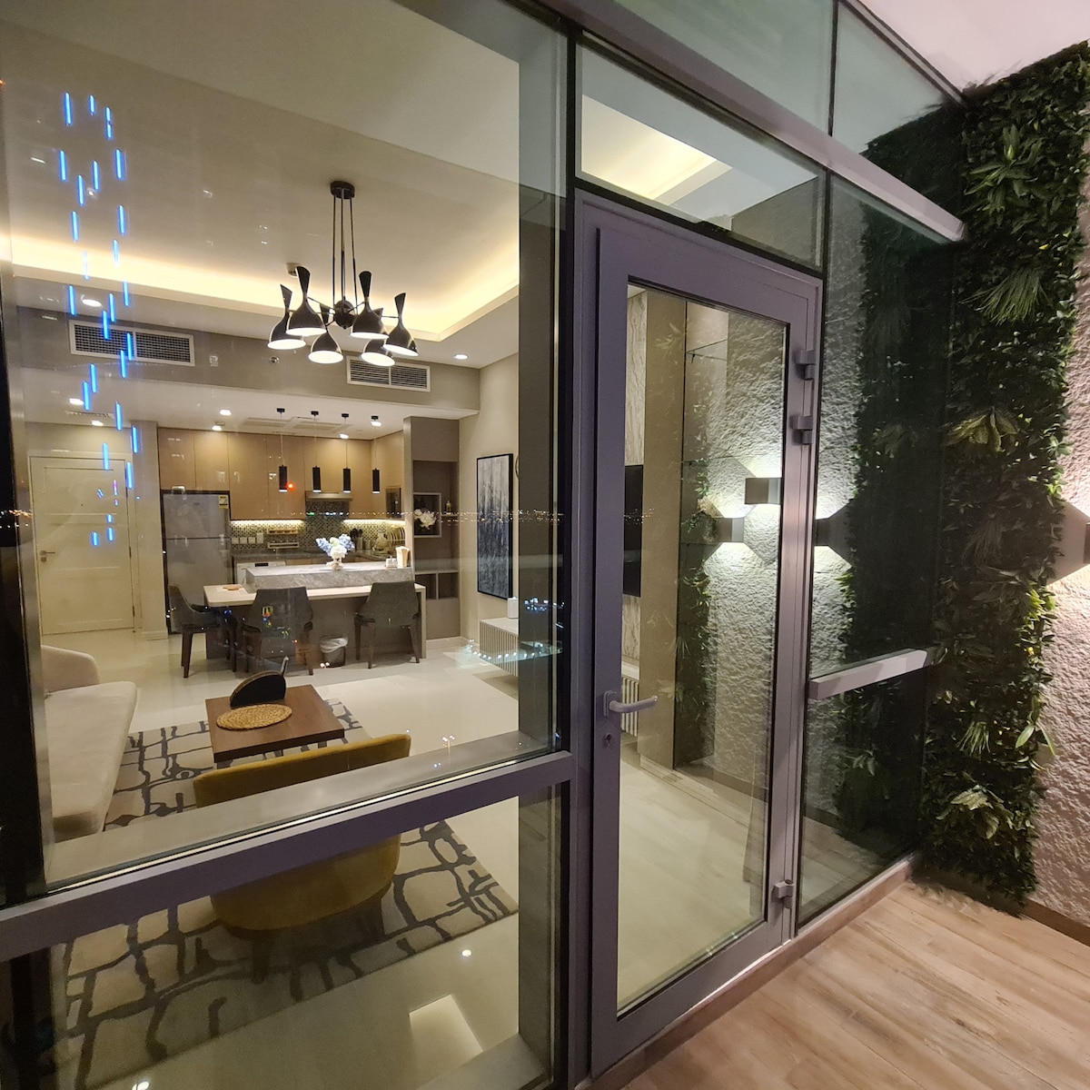 The entrance showcases a modern glass door framed by textured stone walls and greenery. Inside, a spacious living area is visible, featuring a dining table, stylish lighting, and a comfortable seating arrangement. Warm light creates an inviting ambiance throughout the space.