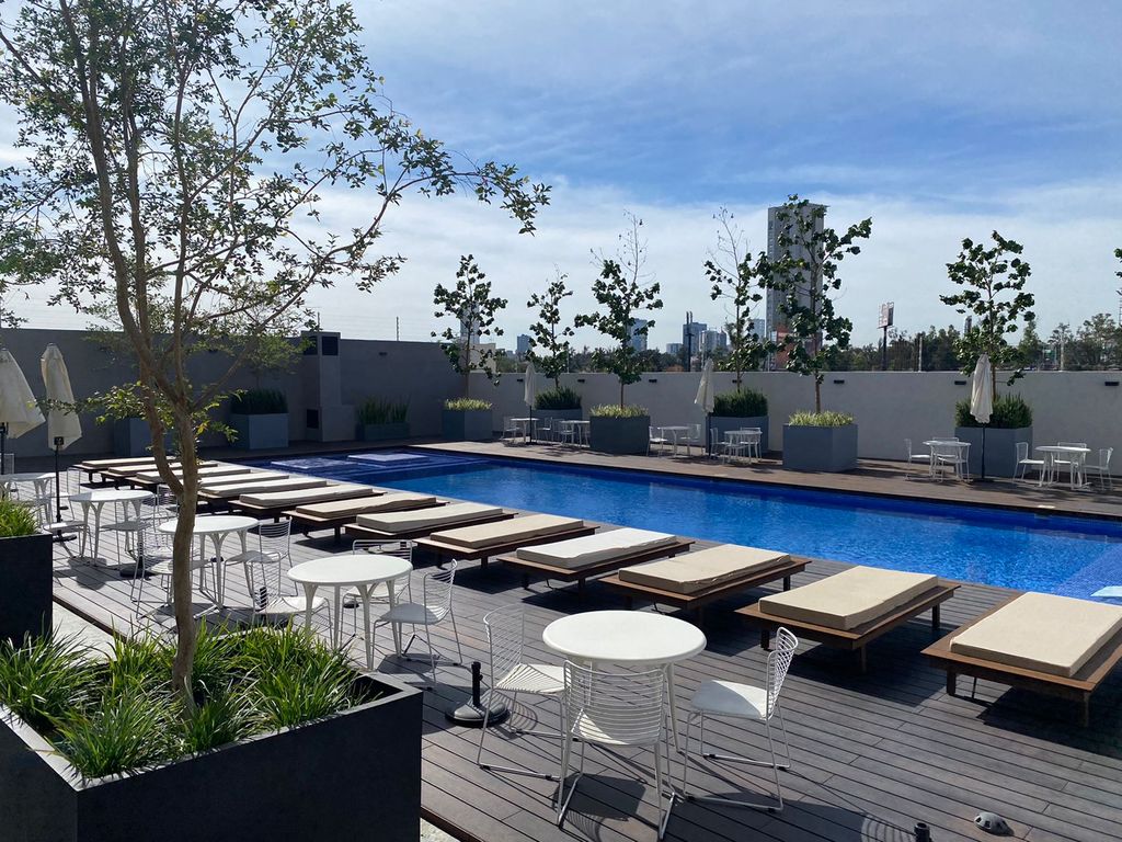 An inviting outdoor pool area features a sparkling blue pool surrounded by sun loungers and patio tables. Lush greenery and decorative planters enhance the space, while several umbrellas provide shaded seating options. The area exudes a tranquil and relaxing atmosphere.
