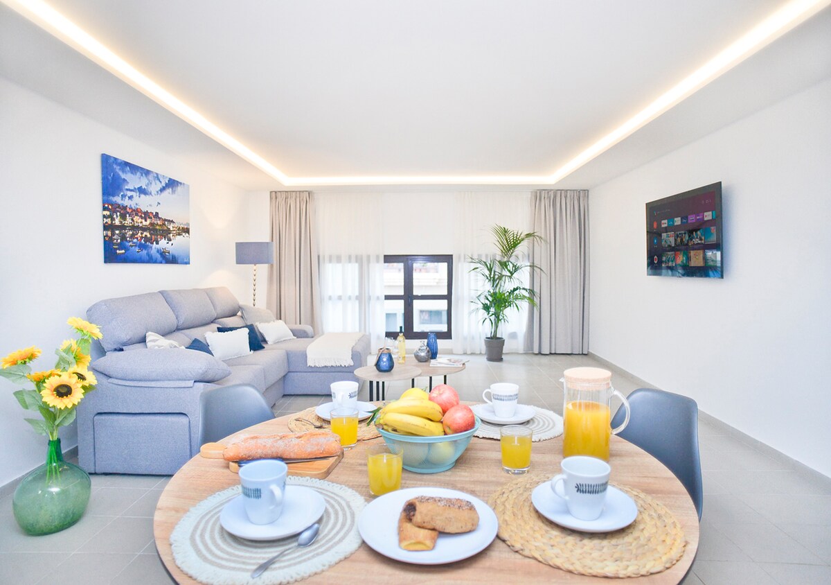 An inviting living area features a comfortable sectional sofa, complemented by light-colored curtains. A round table is set with drinks and fruits, including bananas and apples. A potted plant adds a touch of greenery, while artwork decorates the walls.